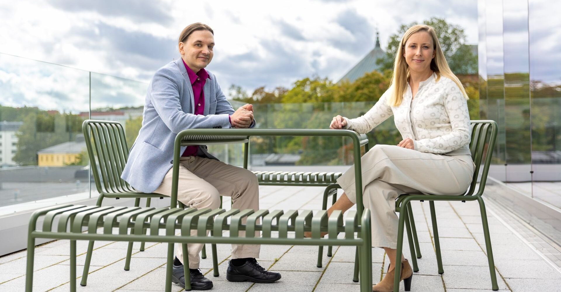 Två av Donnerska institutets doktorander sitter på en takterrass. Ena stipendiaten är en man med mörkt hår, ljusblå kostym och ljusa byxor. Den andra stipendiaten är en kvinna med långt ljust hår och ljusa kläder.
