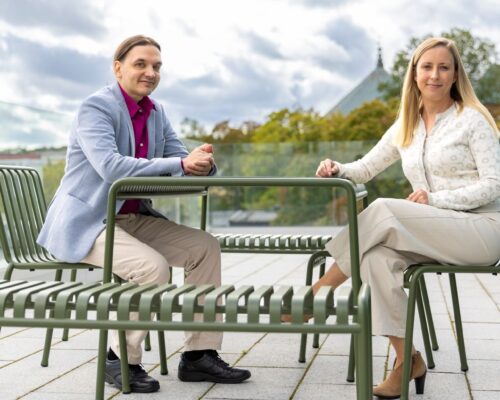 Två av Donnerska institutets doktorander sitter på en takterrass. Ena stipendiaten är en man med mörkt hår, ljusblå kostym och ljusa byxor. Den andra stipendiaten är en kvinna med långt ljust hår och ljusa kläder.